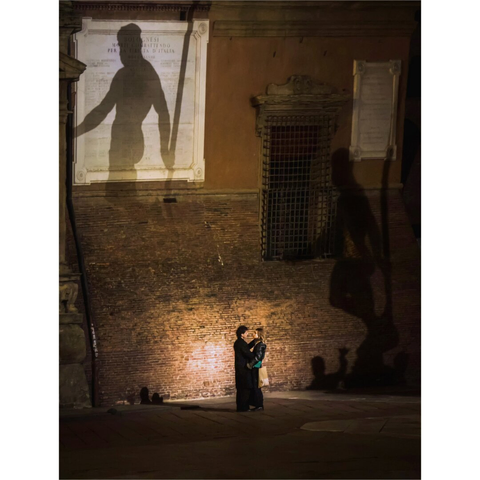 At night shot of a couple is embraced in front of a brick wall (Palazzo d'Accursio)with shadows of statue projected onto it (Neptune). The wall has inscriptions and an old window covered with bars. The lovers are at the bottom of the image illuminated by a light that is out of frame.