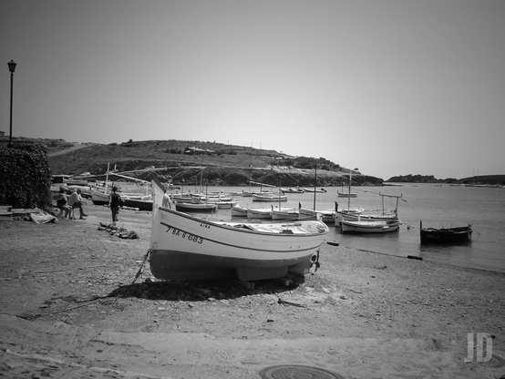 La imagen muestra una pequeña cala con varias barcas de pesca tradicionales, algunas varadas en la orilla y otras flotando en el agua. Al fondo se ven colinas bajas y un cielo despejado. En la orilla, unas pocas personas parecen estar trabajando o conversando junto a las embarcaciones. La escena transmite tranquilidad y un ambiente marinero típico de un pueblo costero.