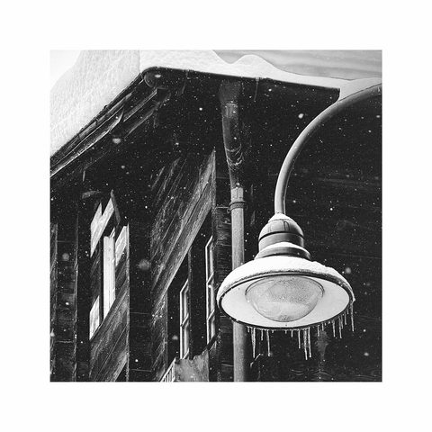a square black and white image showing a detail of some old wooden houses and their roofs. the dark wood builds up a strong contrast to the white window frames and the fresh snow. in the foreground there's a round street lamp with some icicles around the edge