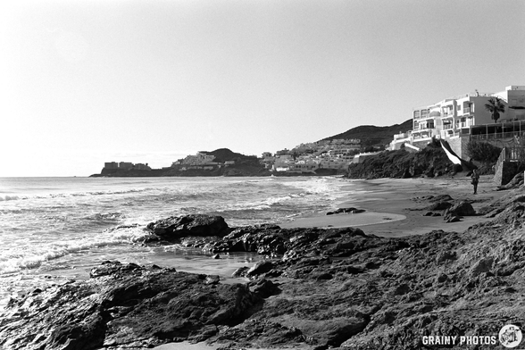 A rocky shoreline with waves lapping against the shore, and a figure walking along the beach. In the background, there are coastal buildings and hills under a clear sky, depicted in black and white.