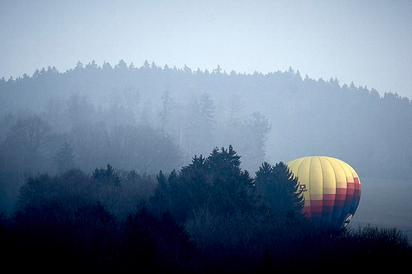 A large, colourful hot air balloon sits nestled among the dark silhouettes of evergreen trees in a misty, wooded landscape. The balloon has a bright yellow top with horizontal stripes of orange, red and blue near its base and displays a small Swiss flag emblem. Thick, blue-toned mist covers the background, partially obscuring a dense forest which rises into a soft, hazy hillside. It is a cold, quiet morning with low, atmospheric lighting. The high contrast between the vibrant balloon and the monochromatic woods evokes a sense of calm.
