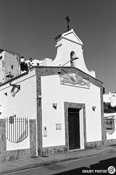 A small, white church with a cross on top, featuring a decorative wrought iron gate and simple architectural details. The setting includes nearby buildings and palm trees, captured in black and white.