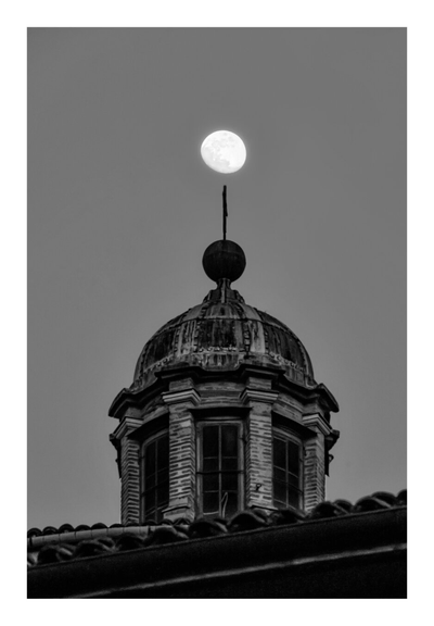 A black and white photograph of a weathered, domed roof with a central spire, positioned against a clear night sky. A full moon is centered above the dome, casting a soft glow. The roof's texture is rough, with visible cracks. The image has a high contrast, emphasizing the moon's brightness against the dark, textured roof.