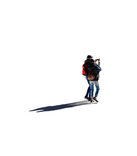 A Photograph of a people standing against a stark white background, casting a long shadows to the left. The people are wearing a black baseball cap, blue jeans, and a jackets. One of them is carrying  a brown leather backpack another another a bag with a red accent. The person on the left with a red backpack is holding up a phone in both hands to take a picture.