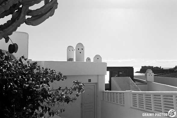 A black and white image of a rooftop scene featuring abstract white structures resembling ghostly figures, surrounded by greenery and distant buildings against a clear sky.