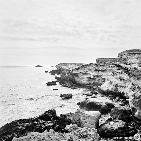 A black and white coastal landscape featuring rugged cliffs by the shore, gentle waves lapping at the rocks, and a distant historic structure overlooking the sea under a cloudy sky.