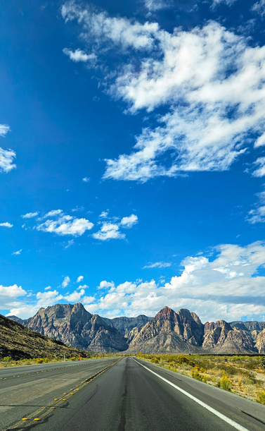 A straight two-lane road with yellow dashed center lines and reflective pavement markers leads toward a dramatic mountain range with jagged red and gray rocky peaks. Sparse desert scrubland lines both sides of the road. The sky dominates the upper two-thirds of the image, a deep cobalt blue filled with scattered white cumulus clouds. The scene is bright and sunny, capturing the stark beauty of the Mojave Desert landscape near Las Vegas, Nevada.