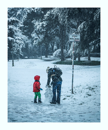 The photo is of a serene winter scene in a park, with a light snowfall creating a soft, ethereal atmosphere. In the foreground, two figures, a child and an adult, are  building a snowman. The child is dressed in a vibrant red jacket and green pants and stands near the partially formed snowman. The adult is facing away from the camera and is shaping the snowman. The surrounding trees are dusted with snow, their branches creating a dark, textured backdrop against the falling flakes.