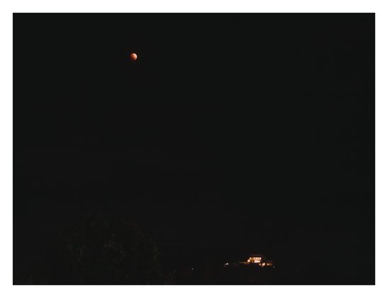 7:19PM / 19:19

High in the upper-left, a small, reddish-orange moon (resembling a "blood moon") stands out against the darkness. At the very bottom, a distant building glows with warm lights, while a dark, indistinct silhouette of a tree is visible on the left.