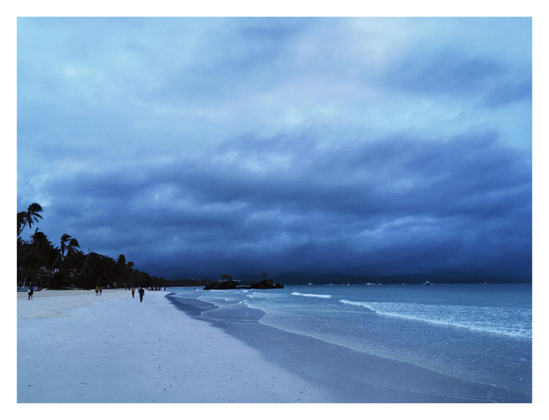 Boracay Island at sunrise, under a sky heavy with low clouds, with blue dawn light washing over everything. A beach of fine white sand with an extremely shallow incline slips beneath the inch-high waves rolling gently to shore. Tiny figures stroll along the sand in the distance. A mass of shadowy palm trees at left. In the shallow water just offshore is a low line of rocks, the site of a religious shrine. Boats dot the horizon, among vague silhouettes of other distant islands.