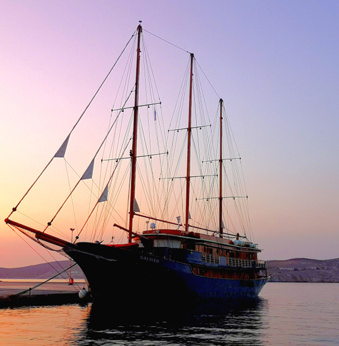 Das Bild zeigt ein grosses Segelschiff/Dreimaster namens "GALILEO" am Abend. Das Schiff hat drei hohen Masten und ist mit einem dunklen blau und rot bemalt. Es ist im Hafen festgemacht, und das Wasser reflektiert die Farben des Abendhimmels, der in sanften rosa- und lila-Tönen erstrahlt. Im Hintergrund ist ein Teil der Insel ersichtlich.
