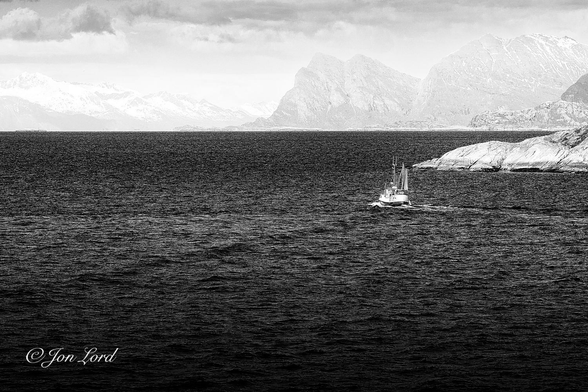 This is a black and white seascape photo in landscape format of a small commercial fishing boat underway with snow covered mountains in the background. Vestfjorden, Norway (2023).

From the base of the photo rising upward to the three quarters up level is a jet black and choppy sea (Sea State 4). There is a clear and crips horizon where the water meets the distant coast ahead, perhaps 10km away. To the right of centre is a small commercial fishing boat, underway, leaving a small white wake behind and heading away from the camera. The view is oblique with the stern and port side in view. The boat is about 10m long, with a black hull, white superstructure and a small sail above the stern. There is a rounded, grey rocky outcrop to the right of the fishing boat. In the far distance, 10km or more away, are two snow covered, steep and rocky mountain ranges. One closer, on the right side and a further more distant range on the left. Above is a sky of broken cloud cover. 