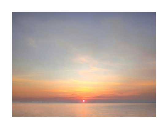 The photo is of a serene sunset over the calm ocean. The sun, a vibrant reddish-orange, sits low on the horizon and casts a warm glow across the sky. The sky is a blend of soft pastels pale oranges, pinks, and blues - with thin, wispy clouds. The water is relatively still, reflecting the colors of the sunset. There is a distant, dark shoreline which is barely visible on the horizon.