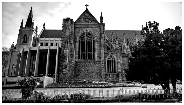 A dramatic black and white architectural photograph of a historic cathedral featuring intricate stonework, a steep roof, and tall, gothic style arched windows. A dark tree stands to the right, and a low metal fence runs across the foreground.