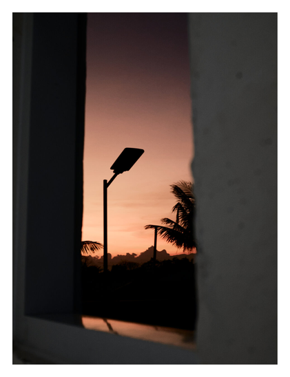 A moody, vertical photograph taken through a dark, narrow window. The view captures a serene twilight scene featuring the sharp silhouettes of a street lamp and palm trees against a soft, gradient sky transitioning from warm peach-orange to deep purple. - Google Gemini 3 Pro Preview
