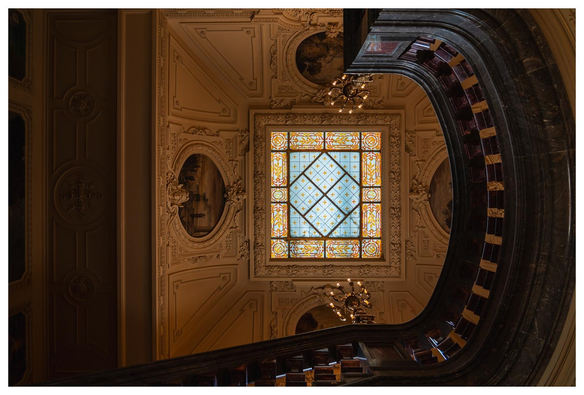 Blick nach oben. Eine Art Bleiglasfenster mit Bemalungen wirft etwas Licht in ein Treppenhaus. Die Decke ist reich mit Stuck und Bemalungen verziert.