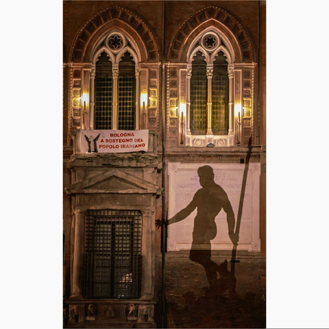 This nighttime photograph is of three windows and a shadow on historic facade of a building in Bologna, Italy (Palazzo d'Accursio) On the top there are two ornate Gothic-style windows with pointed arches and intricate stone which are illuminated by lanterns on either side. Below the left window there is a banner is prominently displayed  "BOLOGNA A SOSTEGNO DEL POPOLO IRANIANO," Bologna supports the Iranian people. 
To the right, a large, towering shadow of Neptune from the fountain is cast against the brick wall, partially overlapping a white commemorative plaque. He is tall, one arm in front of him and one arm behind him holding a trident. On the left beside him is another window.