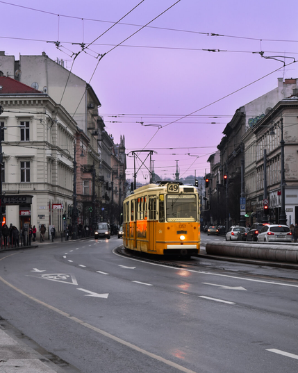 A classic yellow Ganz tram travels along a city street in Budapest at twilight. The tram is illuminated by its headlights and warm interior lights, with the destination sign displaying Line 49.