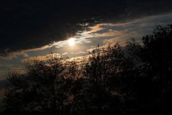 The is seen partially blocked by dark clouds causing the sky to be seen in a dramatic orange glow. The silhouette of trees can be seen along the bottom of the image.