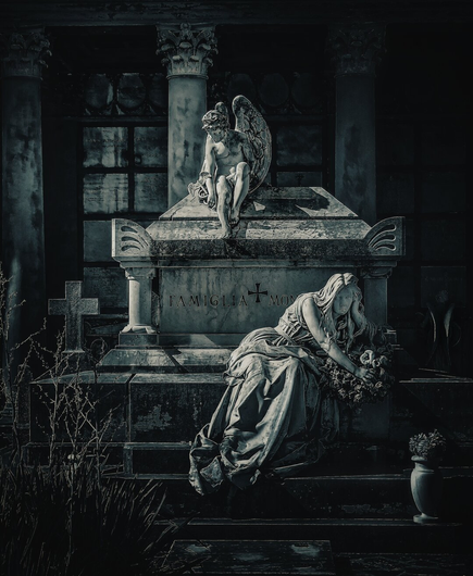 This dark, almost black and white photo was taken nel cimitero monumentale La Certosa di Bologna. It shows a weathered stone tomb inscribed with the name of the family. A sorrowful angel sits on top the monument, looking down at a life- sized statue of a grieving woman who reclines against the base. There are classical columns and a dark, shadowy background which frame the tomb.