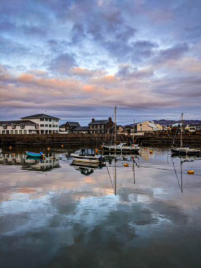 Several small boats moored at Porthmadog Harbour in Wales at dusk, with the cloudy, sunset-tinted sky reflected in the calm water.