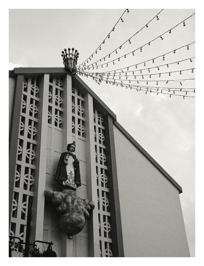 A low-angle, black-and-white photograph of a church facade. The building features strong vertical lines with geometric latticework patterns. Centrally mounted on the wall is a statue of a religious figure standing on a sculpted cloud base. At the peak of the roof sits a crown-like structure, from which multiple strings of festive light bulbs drape outwards against the sky.