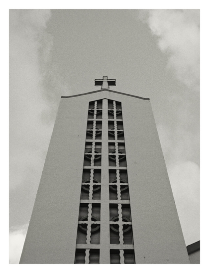 A low-angle, black-and-white photograph of a tall church steeple. The structure rises vertically with plain, expansive walls that taper slightly toward the top, where a simple cross sits at the peak. A central vertical column features a repeating geometric pattern of stylized crosses and circular shapes over louvered vents, creating a striking grid design against the textured, overcast sky.