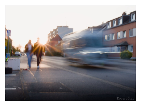 A couple walk hand in hand alongside a road and towards the photographer. The sun is very low in the sky behind them, creating a sunburst over the man's shoulder. A van passes them on the road - the slow shutter speed gives the van's movement a ghostly quality.