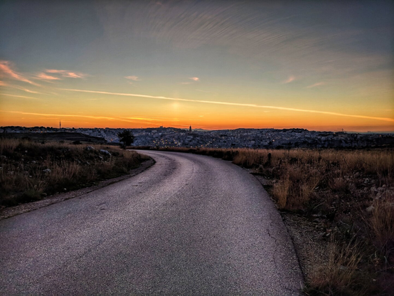 A winding asphalt road cuts through a dry, grassy landscape, leading the eye toward the distant city of Matera, Italy. The scene is bathed in the warm, golden hues of sunset, casting a soft glow over the horizon. The sky is a breathtaking canvas of streaked clouds, transitioning from deep oranges and pinks near the horizon to softer blues above. The cityscape of Matera, with its distinctive architecture and clustered buildings, is silhouetted against the luminous sky, creating a serene and evocative atmosphere.