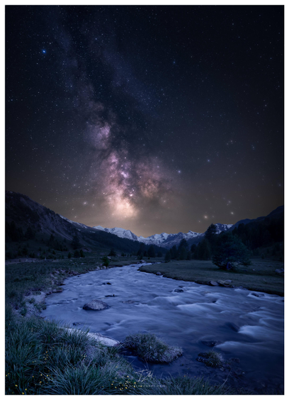 The picture was taken on the bank of a river. The bank is covered with green grass and scattered flowers, which stand out as yellow dots against the otherwise monochrome night scene. The river winds its way through a wide valley dotted with meadows and scattered trees. The Milky Way stretches almost vertically across the valley.
