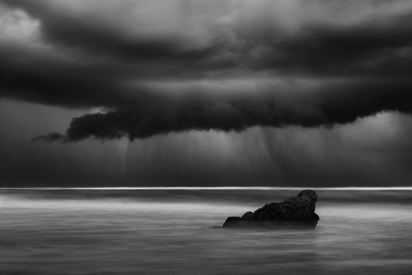 dramatic seascape in black and white. A single triangular rock in the foreground surrounded by slightly smoothed water. A menacing storm cloud in the back with lots of rain coming down.
