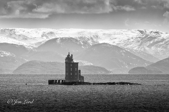 This is a black and white seascape photo of a coastal lighthouse built on a small islet with snow covered mountains in the background. Kjeungskjær fyr, Norge (2023).

The dark, near black sea water covers the lower quarter of the image with small waves of about one metre breaking the surface. Very slightly to the lower left of centre is our octagonal, brick built light house. The lighthouse is on the left side of a low islet or skerry that cannot be much more than a metre above the sea level and is about 50 metres long with a small jetty built on the far left side. The lighthouse or Fyr is octagonal in shape, built from stone and has an octagonal pitched roof with the lantern mounted on the left side above the roof. Beyond the deserted sea is a crisp horizon where the water meets the shore. The land rises with a few areas of rounded mountains with a tall, snow covered mountain ridge in the distant background. Above is a partly cloudy sky with only a few wisps of cirrus clouds. 

Fun Fact - This lighthouse is switched off during the late spring and early summer due to the midnight sun.

https://en.wikipedia.org/wiki/Kjeungskj%C3%A6r_Lighthouse

