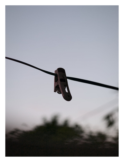 A single dark clothespin hanging on a clothesline. It is silhouetted against a pale, muted sky. In the background below, there are out-of-focus, dark shapes that appear to be trees or bushes. - Google Gemini 3.1 Pro Preview