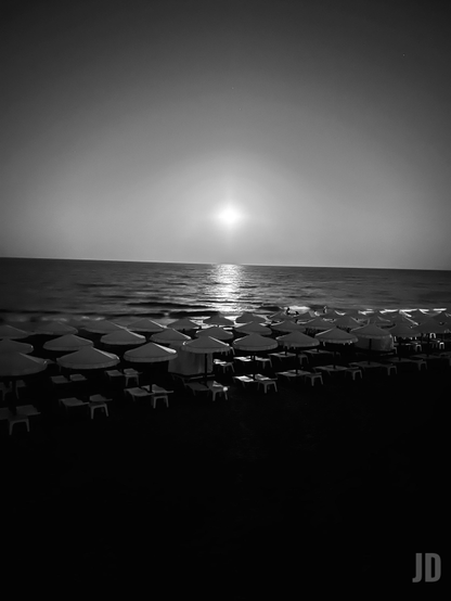 La imagen muestra una playa al anochecer, iluminada por la luz del sol que se refleja en el mar. En primer plano se ven filas de sombrillas y tumbonas vacías, mientras el horizonte y el cielo oscuro crean una atmósfera tranquila y serena.