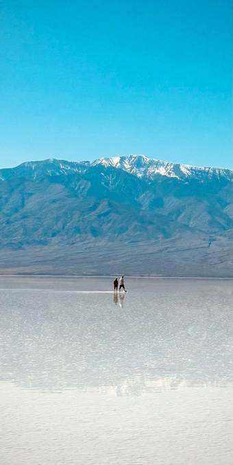 Two people walking on a shallow reflective lake formed on a salt flat with a backdrop of snow-capped mountains under a clear blue sky.
