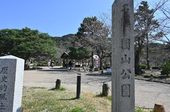 知恩院南門からちょっと入って場所にある円山公園の石碑。奥で桜が咲いている下で傘を指した和服の女性の撮影をしている。/ 2026.03.12 円山公園から