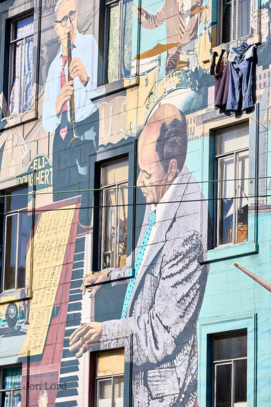 This is a colour street photo in portrait format of the front of a building brightly painted with the figures of various famous jazz musicians playing their instruments on a back drop of local street scenes. Columbus & Broadway, San Francisco (2013).

The facade of the building is viewed obliquely from an angle of about 45º and captures 3 floors above the ground level. The building is a city low-rise apartment block that's neither new nor old. In view are the three floors with three windows on each floor, some open, some closed. Some with curtains drawn, some open. One with laundry drying below the window cill. Stretching upwards from the base to a little above the centre of the photo and dominating the image is the mural of a middle-aged pianist dressed in a jacket and blue tie,  playing the piano with sheet music laid out above the keyboard. Above the pianist and in the upper left corner is a further middle-aged man wearing glasses, shirt and tie and playing the clarinet, Benny Goodman perhaps ? To his right and in the upper right corner is a man playing the drums, his head out of view. The background of the facade in the lower left corner is light blue with the gaps between the musicians being filled with local scenes with street signs, a windmill, sailing boat, car and high-rise in view. 

This is one of the bright and colourful murals that decorate in and around San Francisco's Chinatown. 

