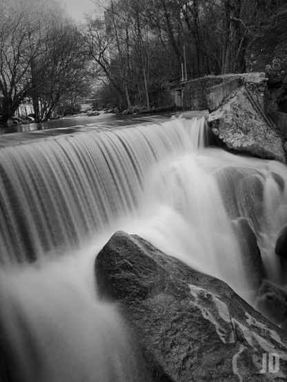 La imagen muestra una cascada de agua que fluye suavemente sobre un muro de piedra, creando un efecto sedoso. En primer plano se observan grandes rocas donde el agua impacta antes de continuar su curso. Al fondo, hay árboles desnudos de hojas, lo que sugiere una estación fría o invernal. La fotografía está en blanco y negro, lo que acentúa los contrastes y la textura del paisaje.
