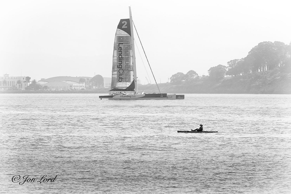 This is a black and white photo in landscape format of a person in a kayak and a more distant sailing boat with part of an island in the hazy background. San Francisco Bay (2015).

The rippled, light grey water of the Bay rises upwards to a little over the half way level. Below and to the right of centre and viewed in profile is a person in a long and slender kayak underway from right to left. In the centre of the photo is a sleek and futuristic Trimaran. The Trimaran is again viewed in profile and is sailing from left to right. The craft has three long sleek hulls of about 30 metres in length and 1 tall mast with the mainsail set and the jib furled. The wind appears to be blowing gently from the viewer towards the boat since it's healing slightly to the left (away from the camera). The boats name: 'Landing Club 2' is written on the side of the three hulls as well as on a large rectangular panel on the mainsail. In the background is part of Yerba Buena Island with the tree lined terrain sloping downwards from the right to left. Where the slope stops is the artificial Treasure Island with some buildings visible on the left. One being the historic Pan Am Clipper Sea Plane terminal and hanger. 

The Trimaran has had numerous names in the past, is at present the 'IDEC Sport' and has broke a vast amount of records for speed, onetime reaching 37 knots.

https://en.wikipedia.org/wiki/IDEC_Sport
https://en.wikipedia.org/wiki/Treasure_Island,_San_Fran