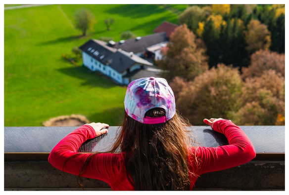 Wir stehen auf einem Aussichtsturm und blicken nach unten auf Wiesen, Wälder und ein Gehöft. Der Fokus ist auf einem von hinten sichtbaren Mädchen mit rotem Pulli und Schirmmütze, das nach unten blickt.