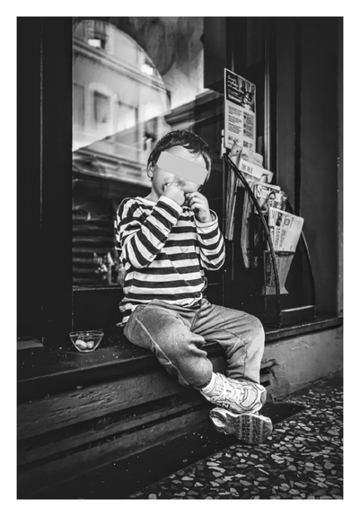 A young child sits on a step in front of a glass door while eating something (taralli) from their hands. They are wearing a striped shirt and jeans. Their face is obscured by a gray rectangle. Next to them on the step is a small glass bowl which has more taralli in it. There are magazines visible in a rack behind them.