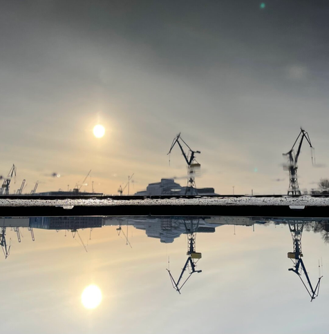 Das Foto zeigt eine Wasserspiegelung mit Sonne und zwei Hafenkränen