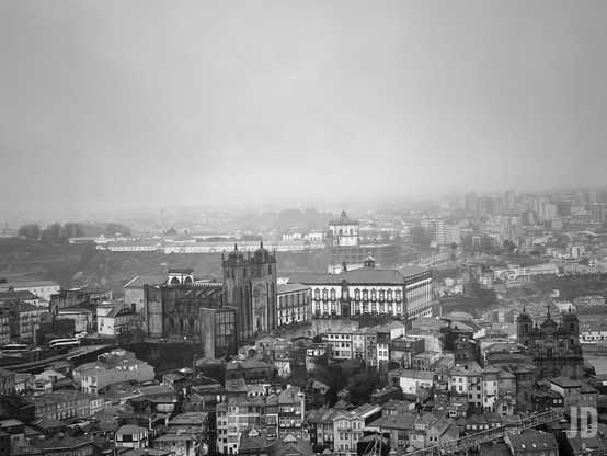Esta es una fotografía en blanco y negro que captura una vista panorámica de la ciudad de Oporto, Portugal, bajo un cielo denso y neblinoso.
La imagen destaca varios elementos icónicos:
• Arquitectura Histórica: En el centro sobresale la Catedral de la Sé de Oporto, con sus torres gemelas de aspecto fortificado, junto al Palacio Episcopal.
• Trama Urbana: Se aprecia la densidad de los tejados tradicionales y edificios estrechos que caracterizan el casco antiguo (Ribeira).
• Atmósfera: La niebla o calima en el fondo difumina los edificios modernos y las colinas distantes, dándole a la escena un aire melancólico y atemporal.
• Composición: La toma está realizada desde un punto elevado, desde la Torre de los Clérigos, permitiendo ver la transición entre la arquitectura medieval y la más contemporánea al fondo.