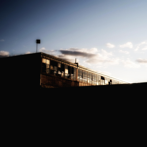 Looking towards the top level of an abandoned industrial building broken windows can be seen in the glow of the evening light. The setting sun causes the top part of the building to glow orange and the bottom half is completely black in shadows. Some soft clouds can also be seen.