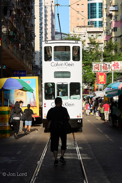 This is a colour street photo in landscape format of a narrow city street with a single pair of tram track laid in the road with a man walking between the rails and a white double-deck tram ahead of the man. North Point, Hong Kong (2014).

From the base of the centre of the photo are two shiny rails set into the roads tarmac surface and stretch away from the camera converging slightly with increasing distance. A little way ahead of the camera, say 3m, is a man, seen from the back, walking away from the viewer and seen in silhouette. 3m ahead of the man is the double-deck, brilliant white, electric tram, in motion (slowly) and away from the man. The tram has the words advertising 'Calvin Klein' in large black letters on each deck as well as the tram's destination 'North Point' on a green roller blind built into the tram. Above the tram is a tall and long single pole connecting the tram to the overhead electric power cable. On either side of the narrow street are market stalls together with people shopping, the closer ones in shadow, the more distant illuminated by the bright and low setting sun. Above the stalls are medium-rise, mixed use buildings, the ones on the left in shadow, one the right: brightly lit. Beyond the tram and on the right are some overhead street signs with large Chinese characters written horizontally across. At the top of the image between the buildings is a narrow strip of clear blue sky. 