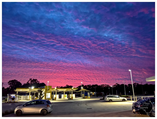 A wide angle landscape photograph capturing a dramatic sunset over a car wash and petrol station in Adelaide. The sky is filled with a dense pattern of altocumulus clouds, creating a rippled texture.

These clouds transition from a deep, glowing magenta and pink near the horizon to a dark violet and navy blue at the top of the frame. In the foreground, a well lit service station features several white washing bays with yellow accents and peaked canopy roofs.

A silver hatchback is parked in the lower left, while a white station wagon and a dark car are visible on the right. Tall outdoor floodlights illuminate the asphalt ground, casting a warm glow that contrasts with the cool, dark silhouette of a line of gum trees in the background.

A small watermark in the bottom right corner features a map of Australia and photography branding.