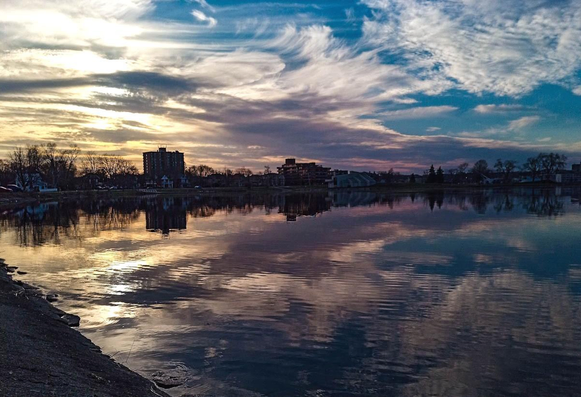 A dramatic sky at sunset overlooking the water of a small lake. Clouds of various sizes fill the sky and are aglow with the setting sun. The calm waters reflect the clouds and the silhouette of the the shoreline covered in trees and buildings can also be seen. 