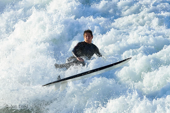 This is a brightly lit, colour water sports photo in landscape format of a surfer having just fallen from his surfboard and in a maelstrom of foaming surf. Hermosa Beach, California (2014).

Filling the entire photo is a mass of brilliant white foaming surf from a recently broken, large Pacific wave. In the centre of the image is a young to middle-aged man, viewed from the front and facing the camera. He is in a near sitting position, having been ejected by the force of nature from his surfboard. Our surfer has dark, slightly spiky hair, his mouth clamped shut and is wearing a black wetsuit. The right side of his face is brightly lit from the still low, morning sun. In front of him and at right angles to him and the camera, is his surfboard: airborne and viewed edge-on with the two shark-like white fins at the rear of the board. 