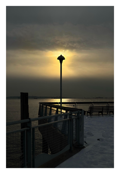 An overcast winter afternoon on the Brooklyn waterfront. The sun glows through a thick veil of clouds. Directly in front of it is a lamppost on a snow-covered pier, standing in silhouette and aligned so that rays of sunlight appear to radiate out from its head. A guardrail bordering the pier recedes from the foreground towards the lamppost in a zigzag line. In the calm waters of the harbor is a column of reflected sunlight that frames the lower part of the lamppost. At left is the silhouette of a wooden pillar rising out of the water. The image is in tones of gray and luminous gold.