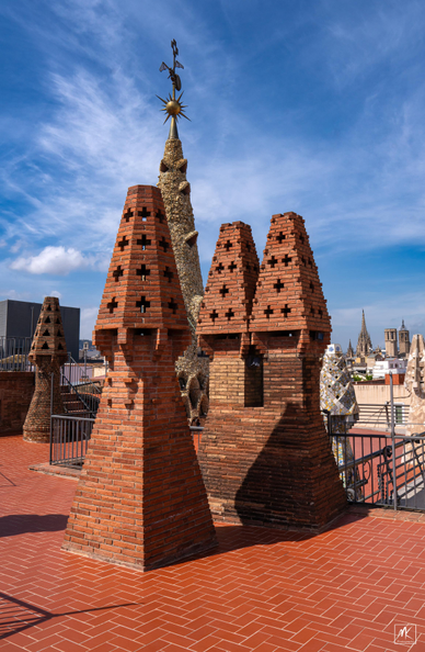Color photo of a group of geometric shaped brick chimneys on a rooftop, with the city of Barcelona in the background. 